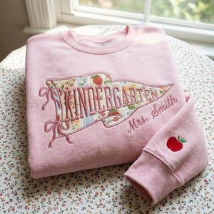May include: A light pink crewneck sweatshirt featuring a large pennant-shaped applique with a floral and strawberry pattern. The text "KINDERGARTEN" is embroidered in pink, with "Mrs. Smith" below it. A small red apple is embroidered on the sleeve.