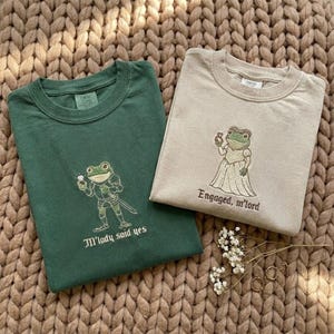 May include: Two crew neck sweatshirts, one green and one beige, with frog illustrations. The green sweatshirt features a frog knight with the text "M'lady said yes." The beige sweatshirt shows a frog bride with the text "Engaged, m'lord."