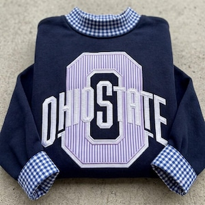 May include: A navy blue long-sleeve sweatshirt featuring a mock neck collar with a blue and white gingham pattern. The cuffs also feature the same gingham pattern. The front displays an embroidered Ohio State logo with white lettering.