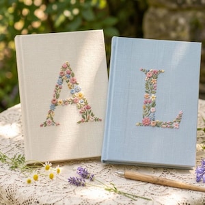 May include: Two hardcover books with fabric covers, one cream and one light blue. Each book features a large, embroidered letter on the front cover, 'A' on the cream book and 'L' on the blue book, both decorated with colorful floral patterns.