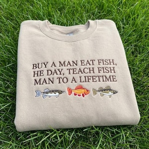 May include: A tan sweatshirt embroidered with the phrase "BUY A MAN EAT FISH, HE DAY, TEACH FISH MAN TO A LIFETIME." Below the text are embroidered images of three different fish in shades of blue, orange, and green.