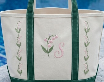 May include: Cream-colored tote bag with green trim and handles. The bag features embroidered lily of the valley flowers and a pink initial "S". The design includes vertical floral patterns on the sides.