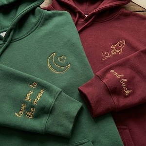 May include: Two hooded sweatshirts, one emerald green and one burgundy. The green hoodie has a gold embroidered crescent moon and heart design, with "love you to the moon" on the sleeve. The burgundy hoodie has a rocket and heart design, with "and back" on the sleeve.