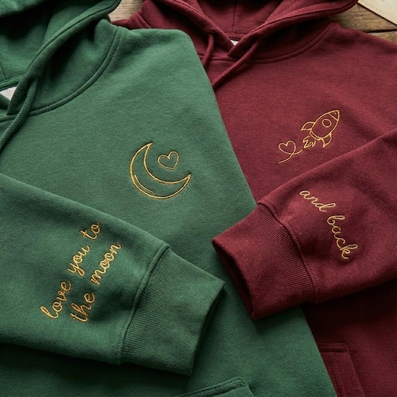 May include: Two hooded sweatshirts, one emerald green and one burgundy. The green hoodie has a gold embroidered crescent moon and heart design, with "love you to the moon" on the sleeve. The burgundy hoodie has a rocket and heart design, with "and back" on the sleeve.