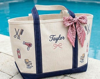 May include: Cream-colored canvas tote bag with navy blue trim and handles. The bag features illustrations of salon tools and the name "Taylor" in script. A pink paisley scarf is tied to the handle. The bag is sitting by a pool.