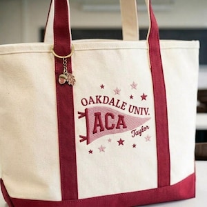 May include: Cream-colored canvas tote bag with burgundy accents, including the handles and base. The bag features embroidered text that reads "Oakdale Univ. ACA Taylor" with star details. A small acorn charm hangs from the handle.