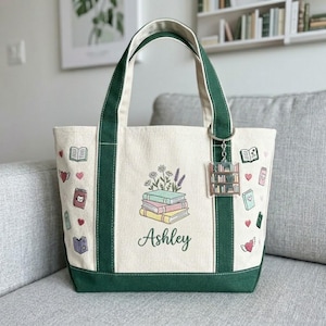 May include: Cream-colored tote bag with green accents, handles, and base. The bag features a stack of books with floral accents and the name "Ashley." Adorned with book and heart illustrations, and a keychain with a bookshelf charm.