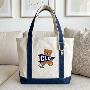 May include: Cream-colored tote bag with navy blue accents and handles. Features an embroidered brown teddy bear holding a pennant with "CLU" and the name "Taylor." A beaded charm hangs from the handle. The bag is sitting on a beige couch.