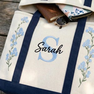 May include: Cream-colored tote bag with navy blue straps and floral embroidery. The bag features the name "Sarah" in script with a light blue "S". Sunglasses, a wallet, and a notebook are visible inside the bag.