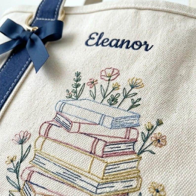 May include: Cream tote bag with navy blue details and a bow. It has an embroidered design of stacked books with colourful flowers and the name "Eleanor" in navy script.