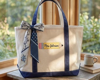 May include: A cream-colored canvas tote bag with navy blue trim and handles. The front features an embroidered yellow pencil graphic with the text Mrs. Johnson. A decorative ribbon with floral and polka dot patterns is tied to the left handle.