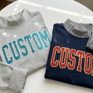 May include: Two long-sleeve shirts with mock neck collars featuring a gray and white gingham pattern. One shirt is heather gray with teal glitter varsity-style lettering reading CUSTOM. The other is navy blue with red glitter lettering reading CUSTOM.