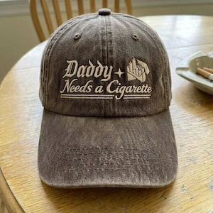 May include: A brown baseball cap with the embroidered text "Daddy Needs a Cigarette" in white script. The cap has a curved brim and a fabric strap closure. The hat is sitting on a wooden surface.