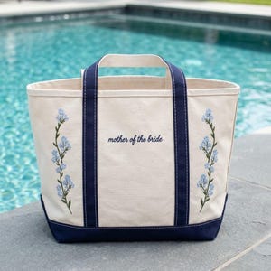May include: A cream-colored canvas tote bag with navy blue handles and a navy blue base. The bag features embroidered light blue floral vines on both sides and the text "mother of the bride" stitched in navy blue thread on the front center.