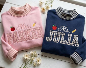 May include: Two long-sleeve sweatshirts with mock necks. The pink sweatshirt features the embroidered text Mrs. PARKER with an apple and pencil. The navy blue sweatshirt features the embroidered text Ms. JULIA with an apple and pencil.