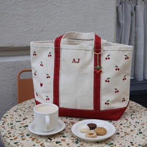 May include: A cream-colored canvas tote bag featuring red cherry embroidery, a single red heart, and the embroidered letters AJ. The bag has red fabric handles and a base, with a gold-toned keychain featuring two red cherries attached to the handle.