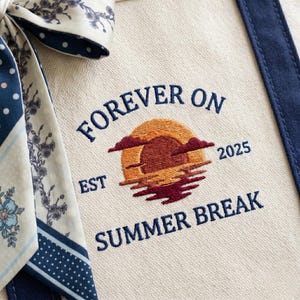 May include: A cream-colored canvas tote bag featuring dark blue embroidered text that reads FOREVER ON SUMMER BREAK with EST 2025. The center design shows an embroidered sunset in orange and maroon hues. A patterned fabric ribbon is tied to the side.