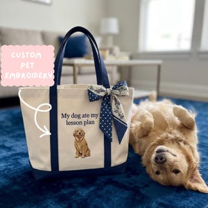 May include: A cream-colored canvas tote bag with navy blue handles and base. The bag features an embroidered golden retriever and the text "My dog ate my lesson plan." A navy blue and white polka dot and floral patterned ribbon is tied to the handle.