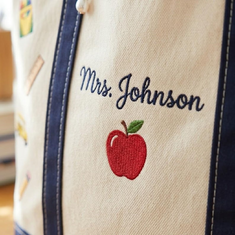 May include: Close-up of a canvas tote bag with navy blue trim and handles. The bag features the embroidered name "Mrs. Johnson" above a red apple with a green leaf. School-themed illustrations are visible.