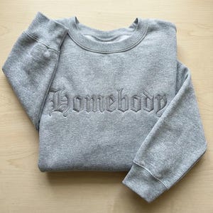 May include: A gray sweatshirt with the word "Homebody" embroidered in a gothic font across the chest. The crew neck sweatshirt is folded neatly on a light wood surface.