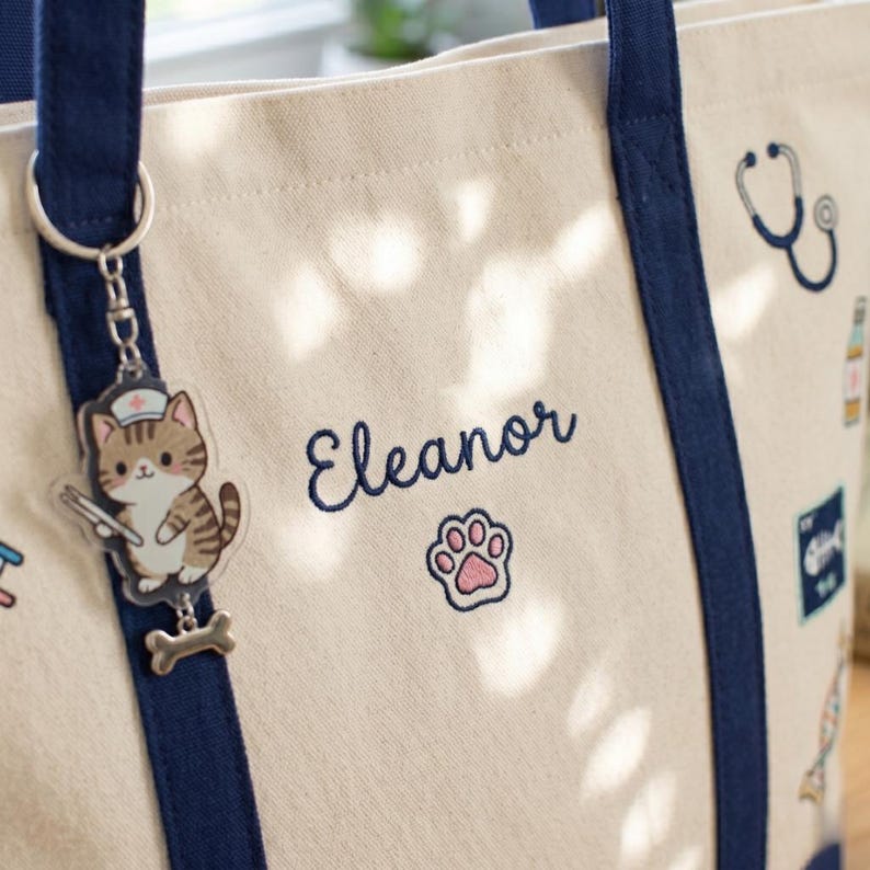 May include: Cream tote bag with navy blue handles and accents. Embroidered with the name "Eleanor" in navy, a pink paw print, and a cat charm. The charm depicts a cat wearing a nurse's hat and holding a medical tool.