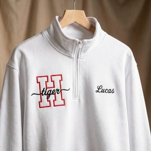 May include: White quarter-zip sweatshirt with a red and black embroidered design. The design includes a large red letter "H" with the word "tiger" in black script. The name "Lucas" is embroidered in black on the right side.
