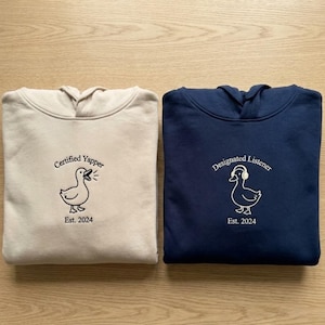 May include: Two hooded sweatshirts, one beige and one navy blue. The beige sweatshirt has a duck graphic with the text "Certified Yapper Est. 2024." The navy blue sweatshirt has a duck graphic with headphones and the text "Designated Listener Est. 2024."