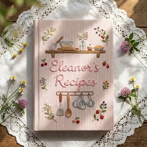 May include: A light pink and white striped recipe book with embroidered kitchen motifs including a shelf with a bowl, whisk, and cupcake, and hanging utensils. The text Eleanor's Recipes is embroidered in pink script across the center.