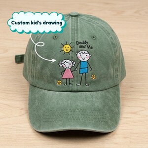 May include: A green baseball cap with a custom kid's drawing. The cap features an embroidered design of a sun, a stick figure of a person, and the text "Daddy and Me". The cap has a curved brim and an adjustable strap.