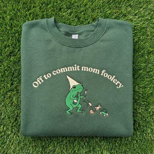 May include: A dark green sweatshirt with the phrase "Off to commit mom foolery" in white arched text. A green frog wearing a hat and holding a bottle and fork is in the center. The frog is surrounded by small cartoon objects.
