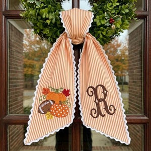 May include: A decorative fabric door hanger featuring orange and white vertical stripes with a white scalloped edge. One side displays an embroidered pumpkin and football design, while the other side features a large, dark brown embroidered letter R.