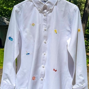 Puede incluir: Una camisa blanca de manga larga con botones, que presenta pequeños motivos bordados de colores dispersos por la tela, incluyendo un sol, una flor, una mariquita y otras formas, terminada con un cuello clásico y puños abotonados.