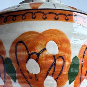 May include: A ceramic bowl with a lid. The white base is decorated with large orange floral designs, white accents, and dark blue outlines. Green leaf shapes and an orange and blue border complete the design. A decorative piece for the home.