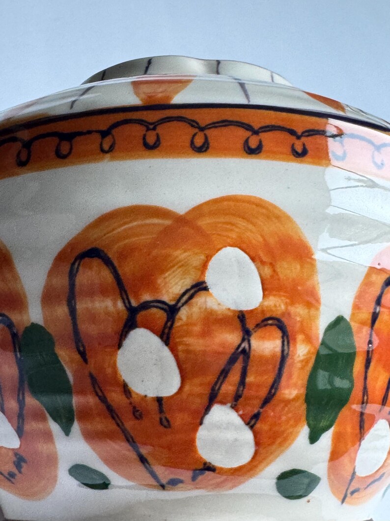 May include: A ceramic bowl with a lid. The white base is decorated with large orange floral designs, white accents, and dark blue outlines. Green leaf shapes and an orange and blue border complete the design. A decorative piece for the home.