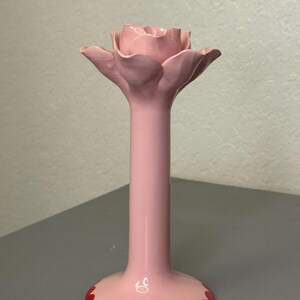 May include: A pink ceramic candle holder in the shape of a rose. The holder features a long stem and a wide base with a scalloped red edge. The rose petals are a lighter shade of pink, creating a delicate aesthetic.