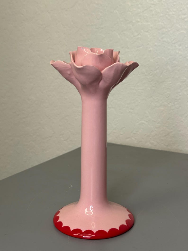 May include: A pink ceramic candle holder in the shape of a rose. The holder features a long stem and a wide base with a scalloped red edge. The rose petals are a lighter shade of pink, creating a delicate aesthetic.