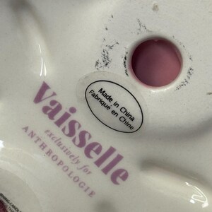 May include: A white ceramic decorative item with a red rim. The base features the text "Vaisselle exclusively for ANTHROPOLOGIE" in purple, along with a sticker that reads "Made in China Fabriqu&eacute; en Chine".