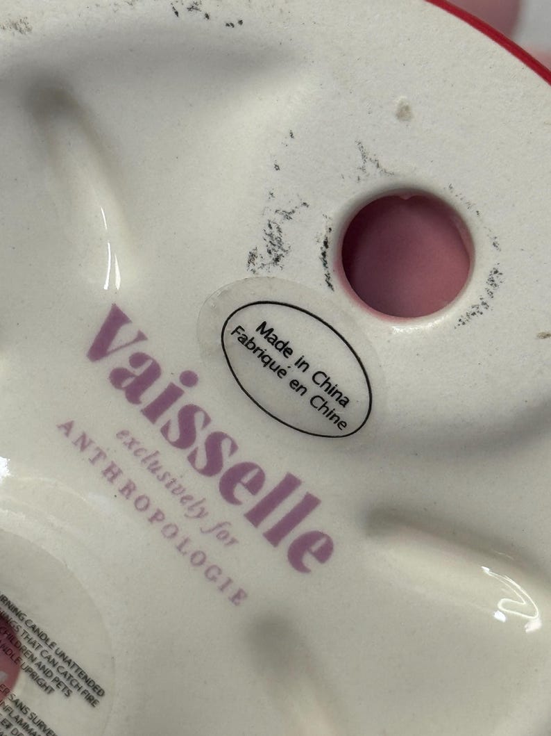 May include: A white ceramic decorative item with a red rim. The base features the text "Vaisselle exclusively for ANTHROPOLOGIE" in purple, along with a sticker that reads "Made in China Fabriqu&eacute; en Chine".