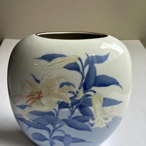 Könnte beinhalten: Eine weiße Keramikvase mit abgerundeter Form, die ein Blumendesign aus Lilien und Blättern in Blau-, Creme- und Rottönen aufweist. Die Vase hat einen goldenen Rand und einen blauen Farbverlauf, was ein dekoratives Stück ergibt.