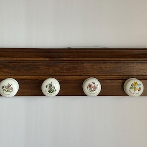 Może przedstawiać: Ciemnobrązowy, drewniany wieszak ścienny z czterema białymi ceramicznymi gałkami. Każda gałka ma inny kwiatowy wzór w różnych kolorach. Wieszak jest zamontowany na białej ścianie.