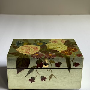 Pode incluir: Uma caixa retangular de madeira verde clara com um design floral. A caixa apresenta um botão dourado e é adornada com flores pintadas em tons de amarelo, rosa e bordô, com folhas verdes. A caixa é provavelmente para joias ou pequenas lembranças.
