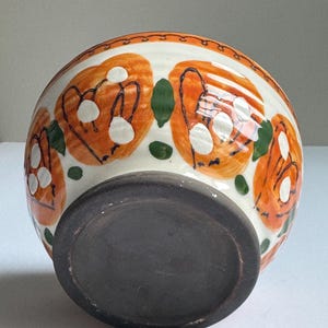 May include: A ceramic bowl with a white exterior, decorated with orange floral patterns and green leaf accents. The rim has a decorative border. The base is dark brown.