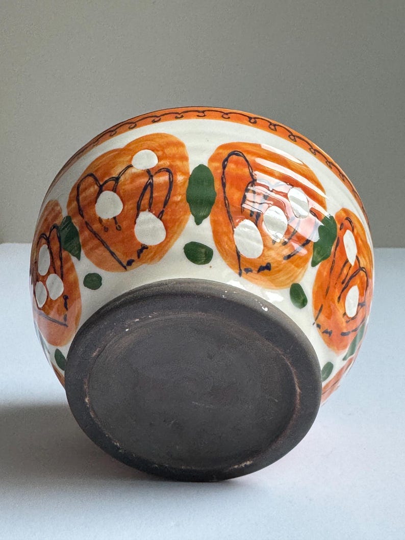 May include: A ceramic bowl with a white exterior, decorated with orange floral patterns and green leaf accents. The rim has a decorative border. The base is dark brown.