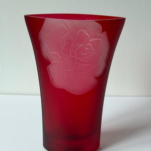 May include: A ruby red glass vase with a frosted rose design. The vase has a flared top and a smooth, curved base. The rose is etched into the glass, creating a delicate contrast.