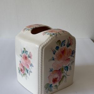 May include: A cream-colored ceramic tissue box cover with a rectangular shape and rounded edges. The exterior features hand-painted floral motifs in shades of pink, peach, and muted green, depicting blooming roses and leaves on the front and top.