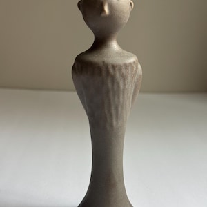 May include: A handcrafted, stoneware figurine in a muted brown hue. The sculpture features a stylized human form with a head, torso, and flared base. The textured surface adds a tactile quality to this unique decorative piece.