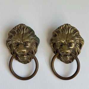May include: Two antique brass lion head door knockers. Each knocker features a detailed lion's face with a ring pull. The brass has an aged patina, adding to the vintage aesthetic. Ideal for adding a touch of classic elegance to any door.