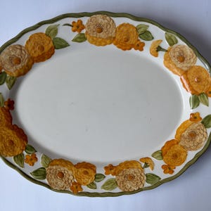 Könnte beinhalten: Eine ovale Servierplatte aus Keramik mit gewelltem Rand und grünem Saum. Die Platte ist mit einem dekorativen Muster aus gemalten gelben und orangefarbenen Blumen mit grünen Blättern auf einem cremefarbenen Hintergrund verziert.