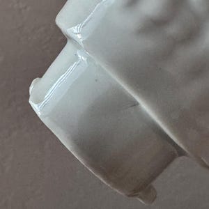 Może przedstawiać: Zbliżenie białego ceramicznego przedmiotu z błyszczącym wykończeniem. Obiekt ma zakrzywioną, teksturowaną powierzchnię i prostokątną podstawę. Oświetlenie tworzy światła i cienie, podkreślając kształt i fakturę przedmiotu.