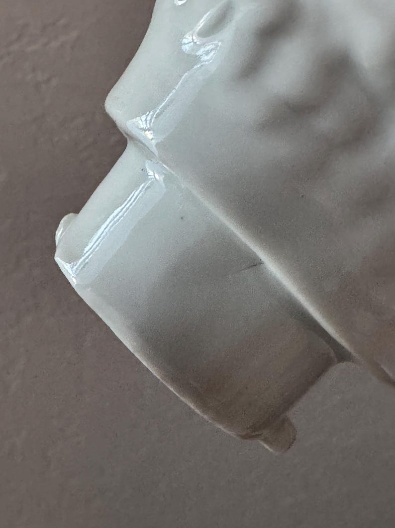 Może przedstawiać: Zbliżenie białego ceramicznego przedmiotu z błyszczącym wykończeniem. Obiekt ma zakrzywioną, teksturowaną powierzchnię i prostokątną podstawę. Oświetlenie tworzy światła i cienie, podkreślając kształt i fakturę przedmiotu.